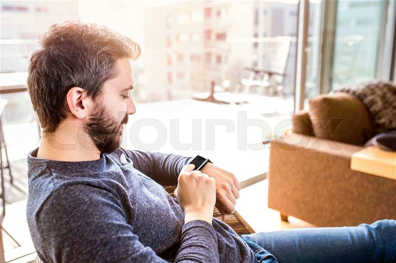 Casual hipster man working from home using smart watch, sitting in living room, stock photo