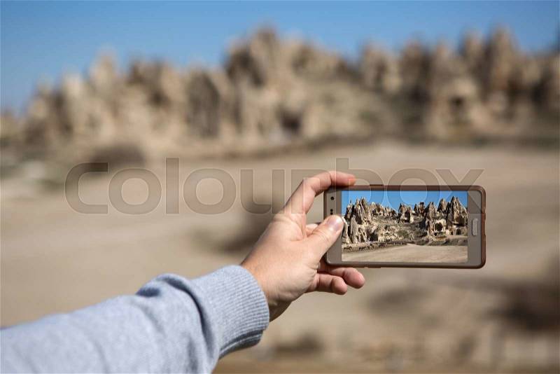 Hand with a phone on the soft background of the rock formation and the blue sky with clouds. On the phone display there is an image of houses carved into rock, stock photo