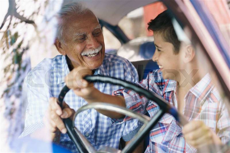 Family and Generation gap. Old grandpa spending time with his grandson and teaching him to drive. The boy holds the volante of a vintage car from the 60s. They both smile happy looking each other, stock photo