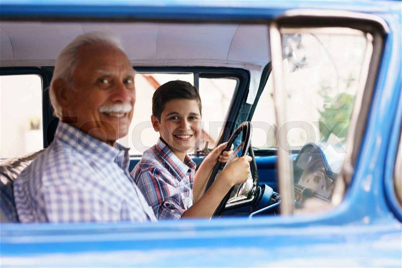 Family and Generation gap. Old grandpa spending time with his grandson. He teaches him to drive. The boy holds the volante of a vintage car from the 60s. They both smile happy looking at camera, stock photo