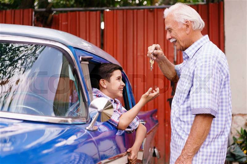 Family and Generation gap. Old grandpa spending time with his grandson. The senior man gives the keys of a vintage car from the 60s to the preteen child sitting inside. They smile happy, stock photo