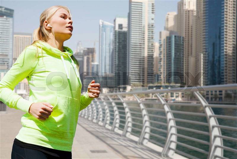 Fitness, sport, people and healthy lifestyle concept - young woman with earphones jogging over dubai city street or waterfront background, stock photo