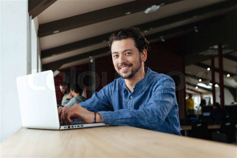 Happy man using laptop computer in office and looking at camera, stock photo