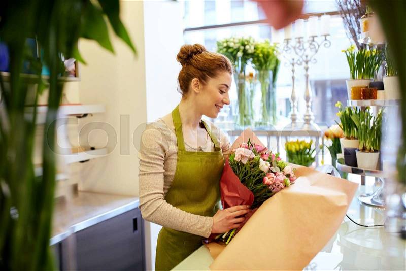 People, business, sale and floristry concept - happy smiling florist woman wrapping bunch into paper at flower shop, stock photo