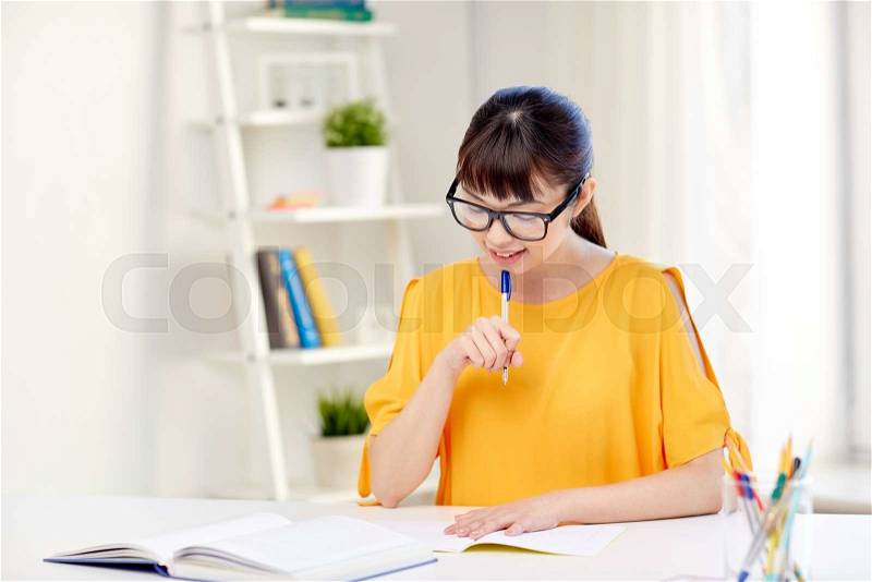 People, education, high school and learning concept - happy asian young woman student in glasses reading book and writing to notepad at home, stock photo