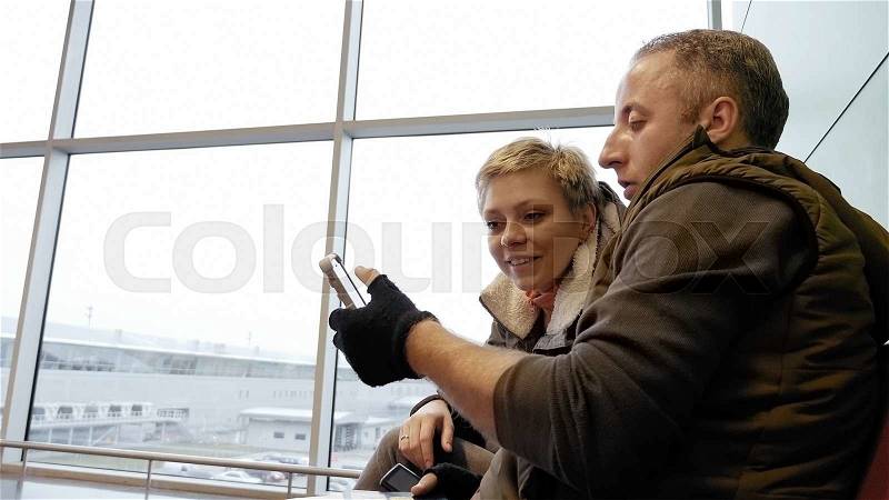 Happy couple or family sitting at the airport terminal and looking at the smartphone screen. Man searches and shows to his girlfriend photos or social media posts. , stock photo