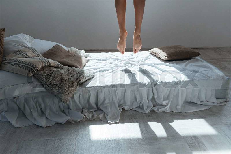 Slim legs of young woman flying in the air above bed in bedroom, stock photo