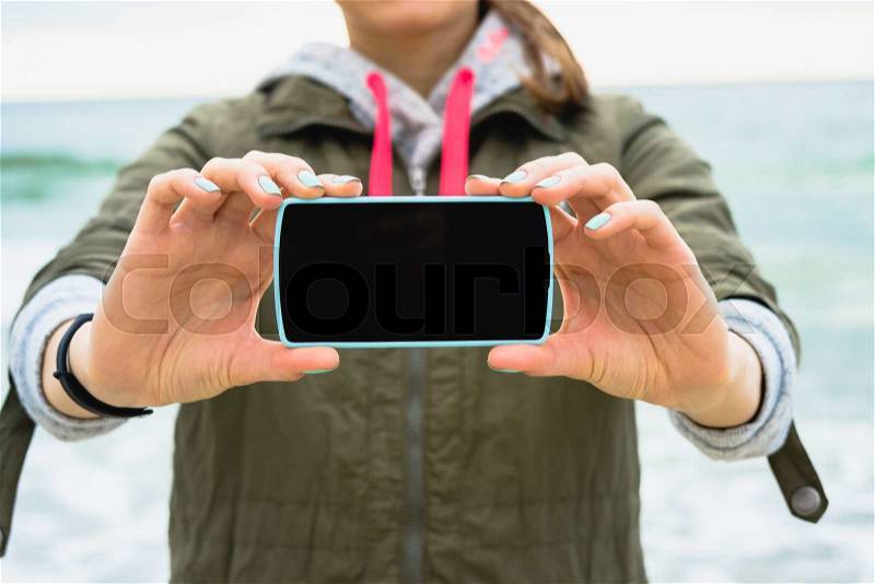 Girl in the green jacket shows a blank phone screen on a background of the sea, stock photo