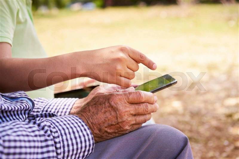 Senior man learning internet and mobile phones: young kid helping his grandpa surfing the web with his new mobile phone. Closeup of both old and young hands touching the screen, stock photo