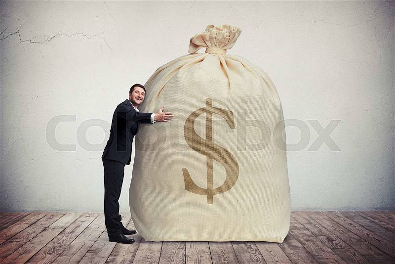 Happy man hugging a big bag of money, grey wall on background, stock photo