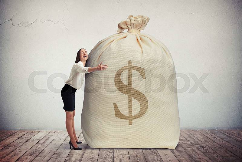 Happy woman hugging a big bag of money, grey wall on background, stock photo