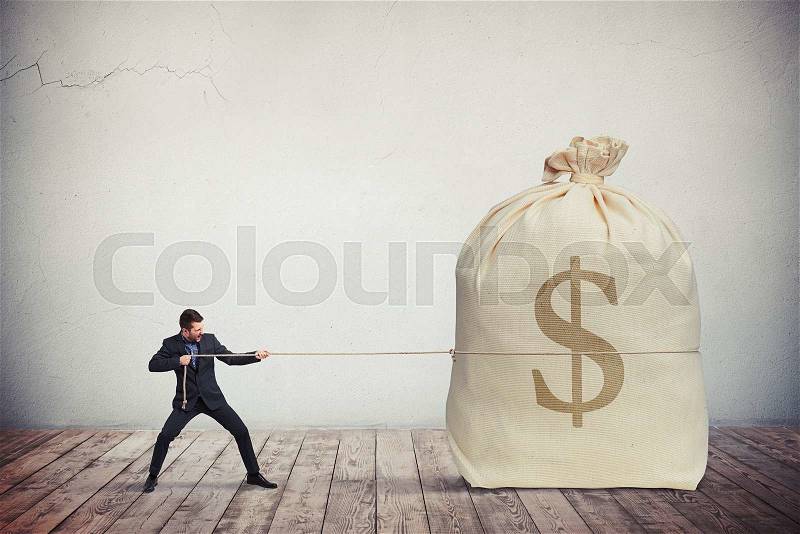 Man pulling on a rope big bag of money, grey wall on background, stock photo