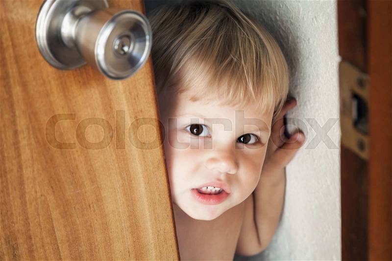 Little Caucasian blond baby girl opens door and looks outside, stock photo
