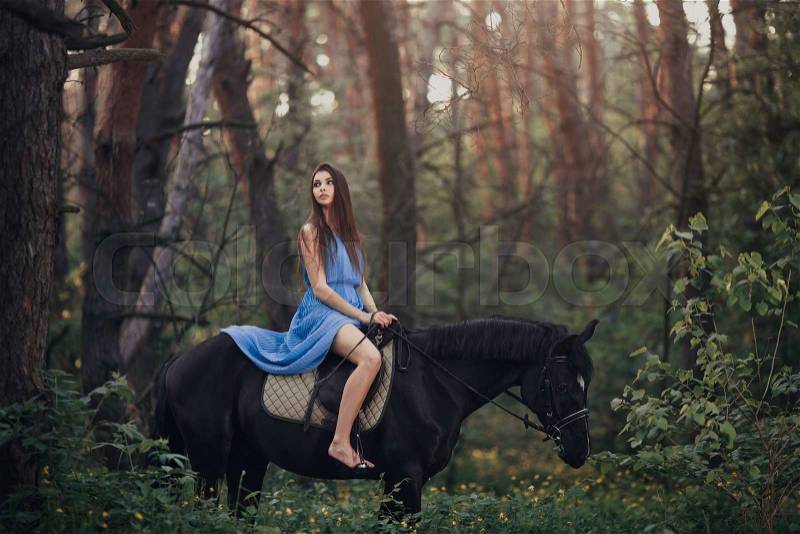 Young beautiful brunette girl in blue dress ride on the black horse in forest, stock photo