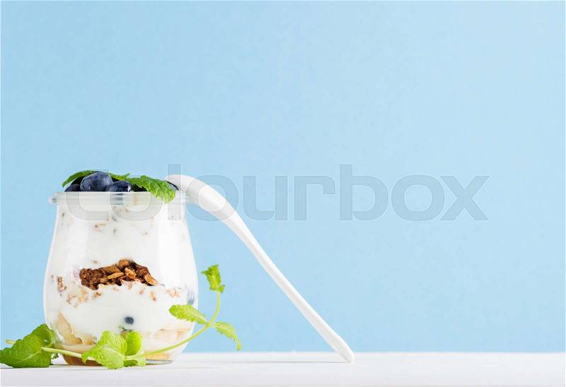 Yogurt oat granola with jam, blueberries and green leaves in glass jar on pastel blue backdrop, copy space, stock photo