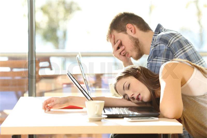 Side view of tired students surrendering to fatigue studying with laptops in a coffee shop with a window in the background and sky outdoors, stock photo