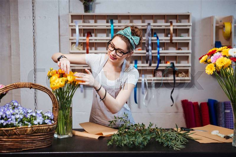 Smiling charming young woman florist taking care of yellow roses in flower shop, stock photo