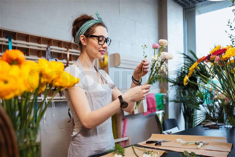 Happy charming young woman florist holding beautiful flowers and making bouquet in shop, stock photo