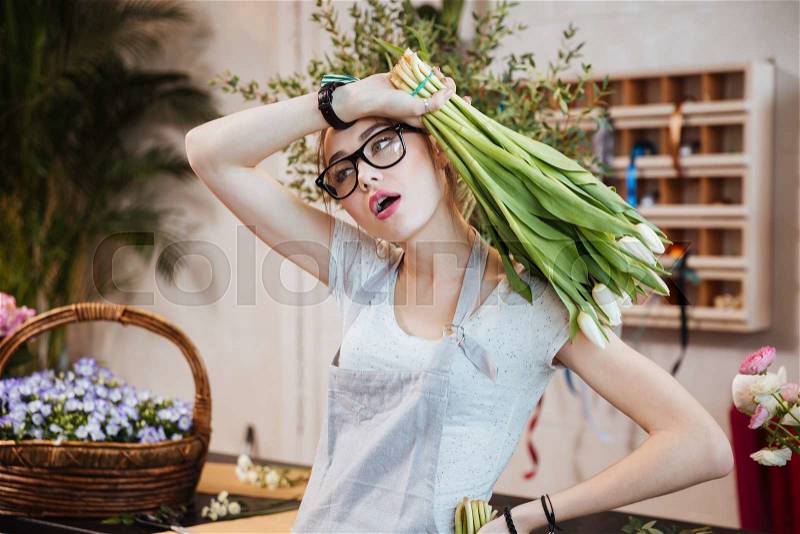 Exhausted pretty young woman florist holding bunch of white tulips and working in flower shop, stock photo