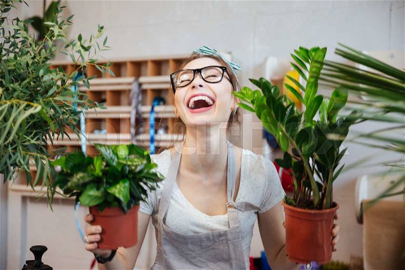 Happy pretty young woman florist holding plants in flowerpots and laughing , stock photo