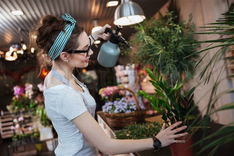 Smiling cute young woman florist standing and watering plants with water sprayer in flower shop, stock photo