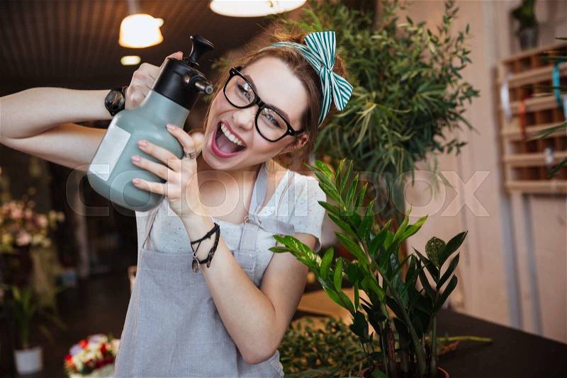 Amusing young woman florist watering flowers ing pointing with water sprayer on you, stock photo