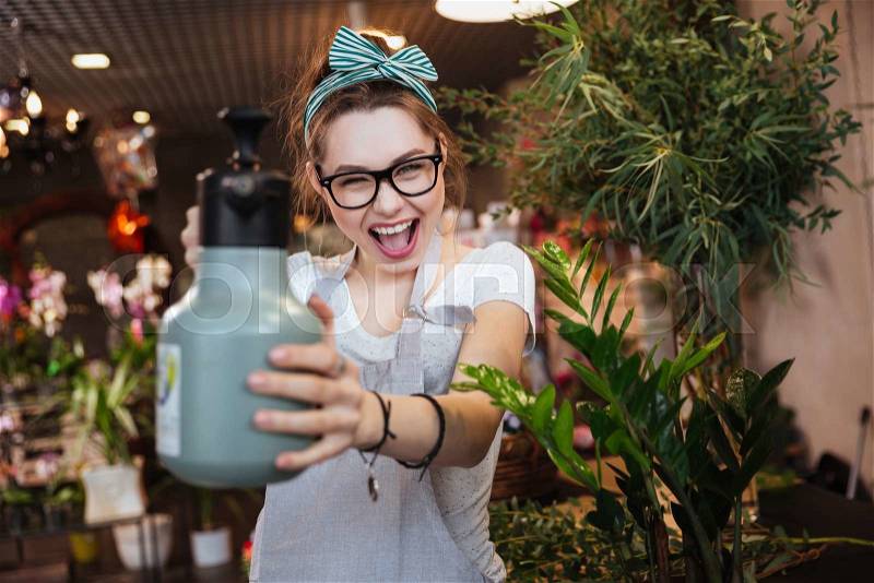 Playful attractive young woman florist pointing with water sprayer on you in flower shop, stock photo