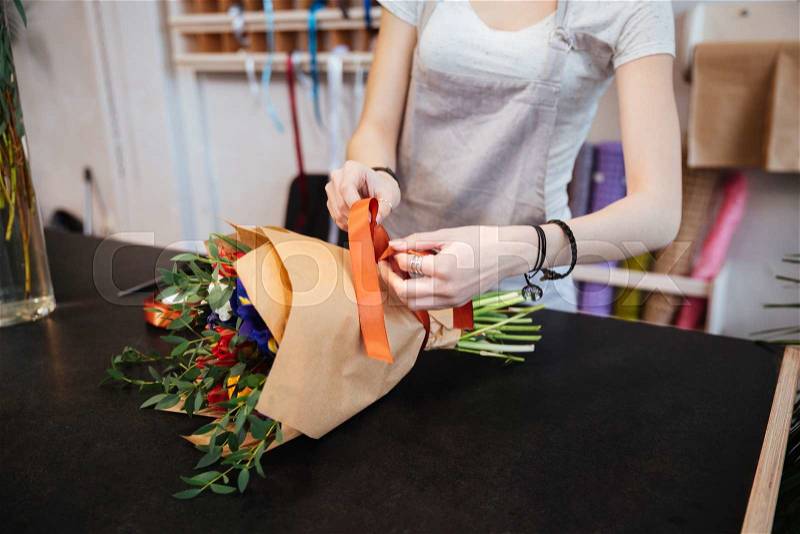 Closeup of hands of young woman florist making bouquet of flowers with red ribbon in shop, stock photo