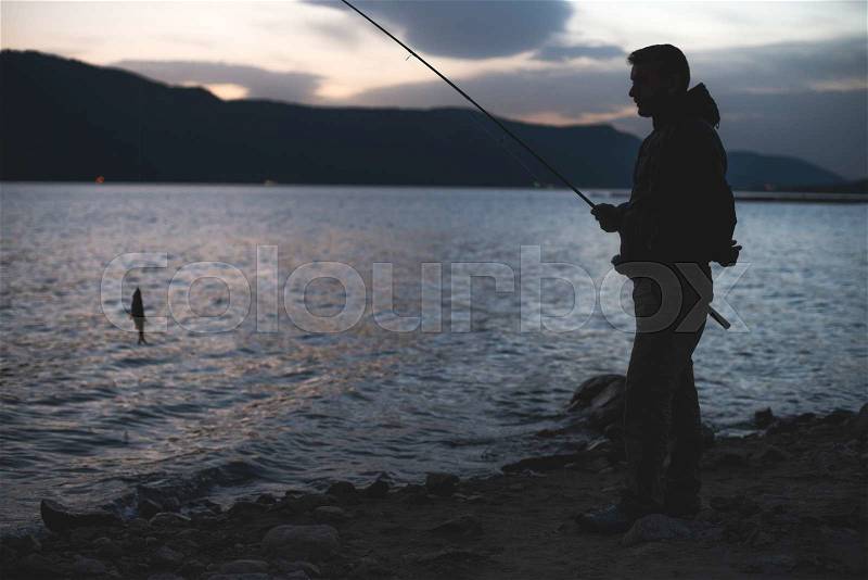 Man fishing on mountain lake. Sunrise low light. Casual clothes, stock photo