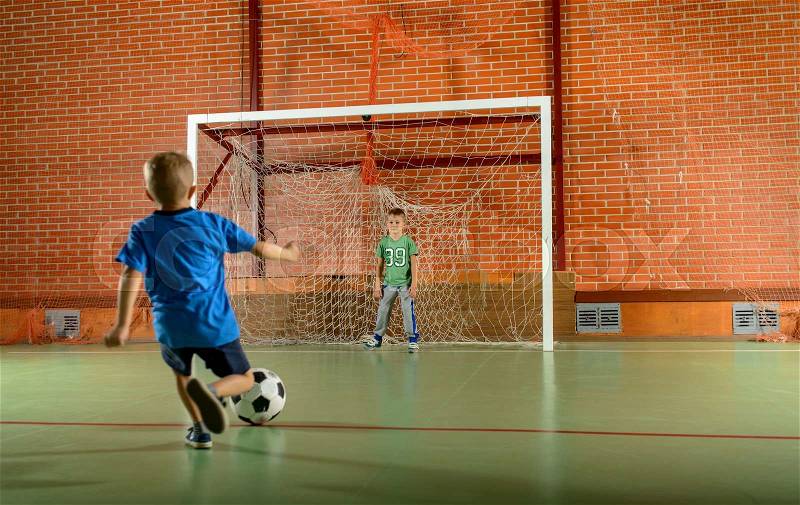 Two young boys playing soccer on an indoor court with one standing in the goalposts as the second prepares to kick for goal, stock photo