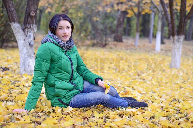 Daydreaming single woman in green winter coat laying back on ground among yellow leaves spread out around her under trees in scenic park, stock photo