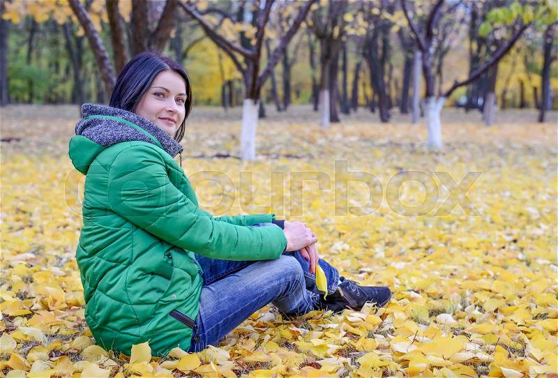 Calm single woman in green winter coat and black hair sitting on ground surrounded by yellow leaves under trees in scenic park, stock photo