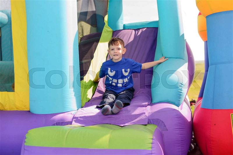 Carefree young boy playing on a bouncy castle exiting a colorful purple slide with his arms outspread and a happy smile, stock photo