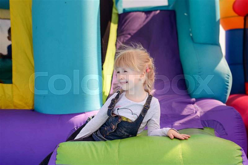 Cute little fair haired girl playing on a colorful bouncy castle at a fairground waving and smiling as she exits the slide, stock photo