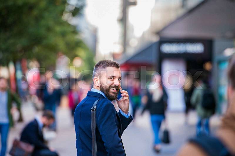 Hipster manager holding a smart phone, making a phone call, walking in the street of London, rear view, stock photo