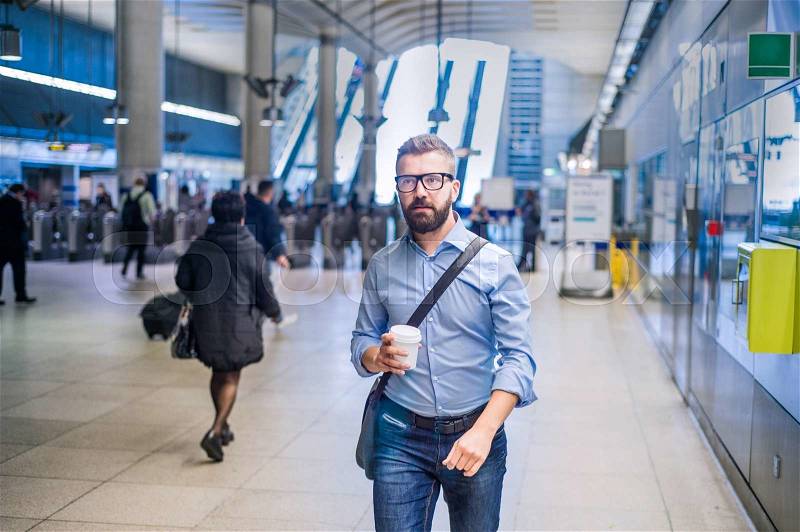 Hipster businessman in blue shirt with coffee cup walking in subway station, stock photo