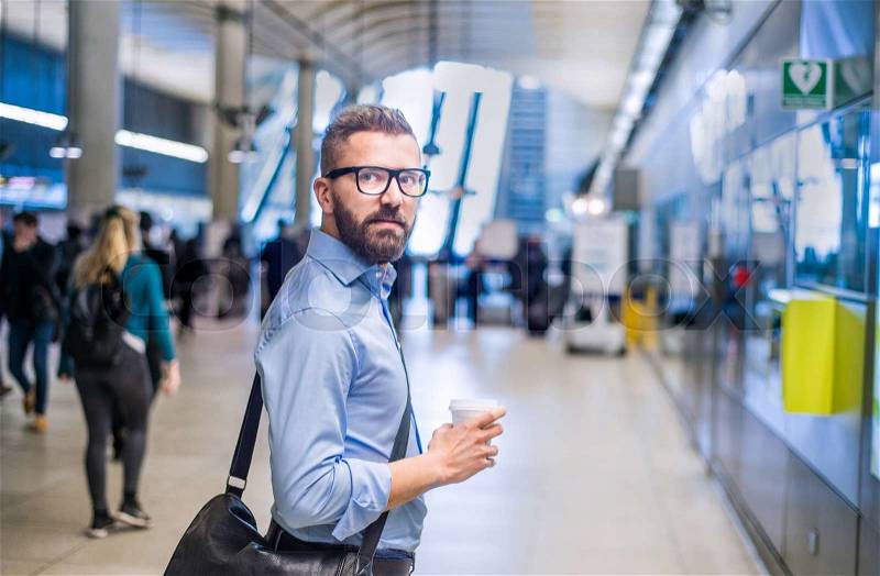 Handsome hipster businessman in blue shirt holding a coffee cup, standing on subway station, stock photo