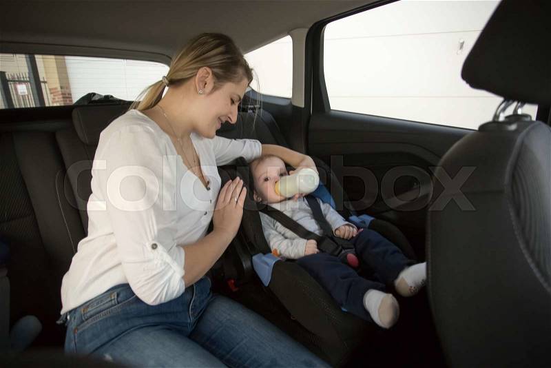 Young mother giving milk in bottle to her little son on car back seat, stock photo