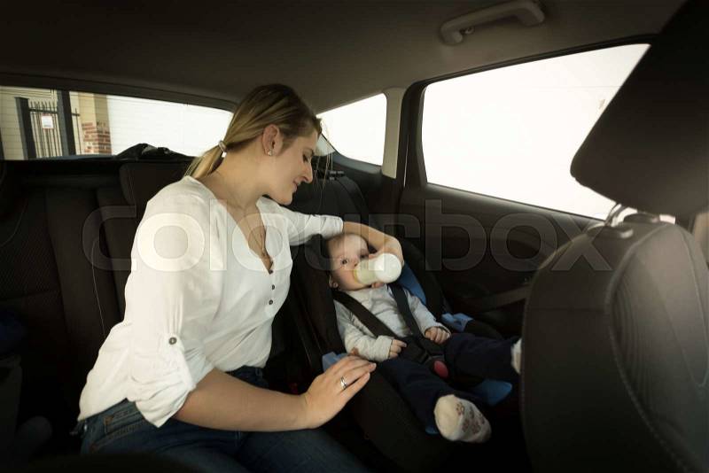 Young mother feeding baby boy with milk on car back seat, stock photo
