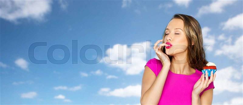 People, holidays, party, junk food and celebration concept - happy young woman in pink dress eating birthday cupcake over blue sky and clouds background, stock photo