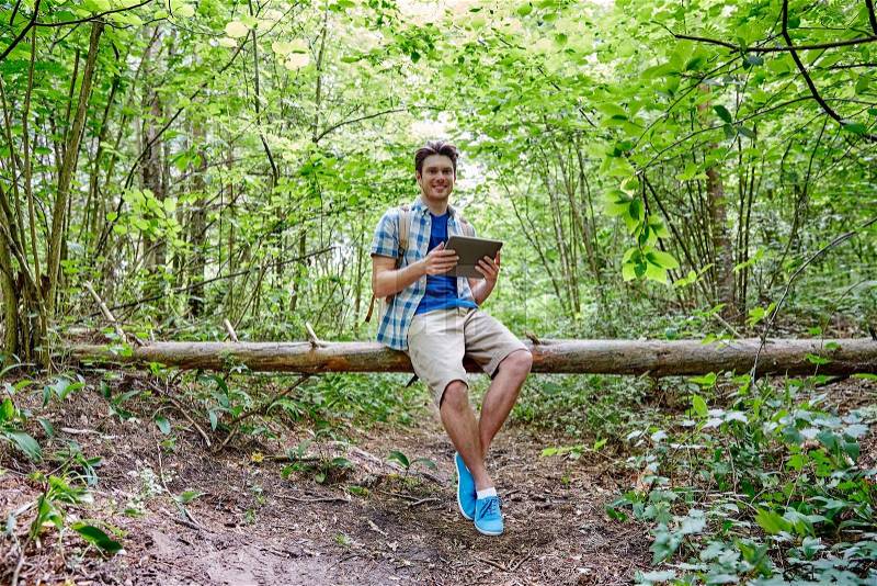 Adventure, travel, tourism, hike and people concept - happy young man with backpack and tablet pc computer sitting on fallen tree trunk in woods, stock photo