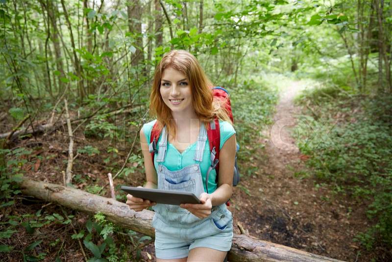 Adventure, travel, tourism, hike and people concept - happy young woman with backpack and tablet pc computer sitting on fallen tree trunk in woods, stock photo