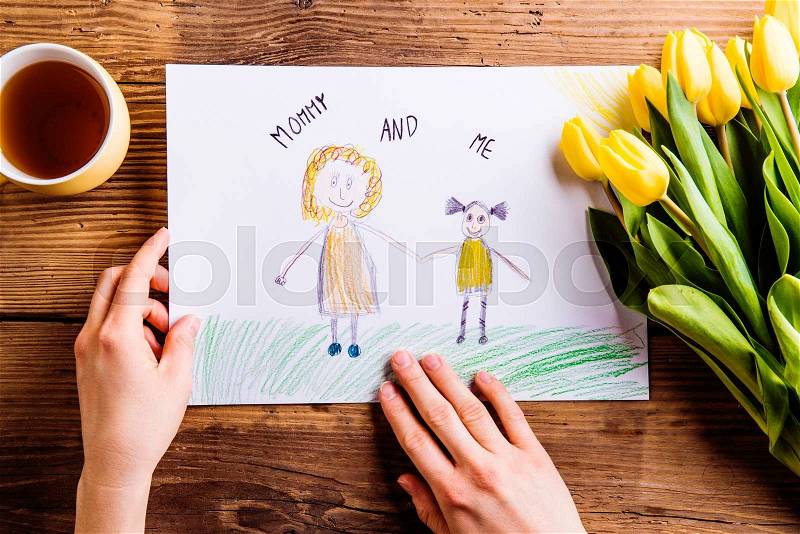 Mothers day composition. Hands of unrecognizable woman holding a drawing of her and her daughter. Bouquet of yellow tulips. Studio shot on wooden background, stock photo