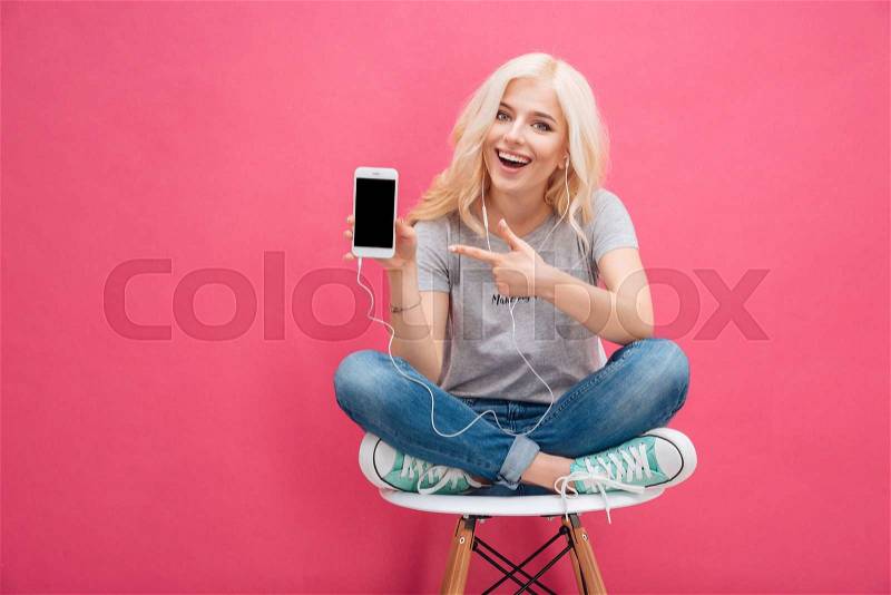 Cheerful woman sitting on the chair and showing blank smartphone screen over pink background, stock photo