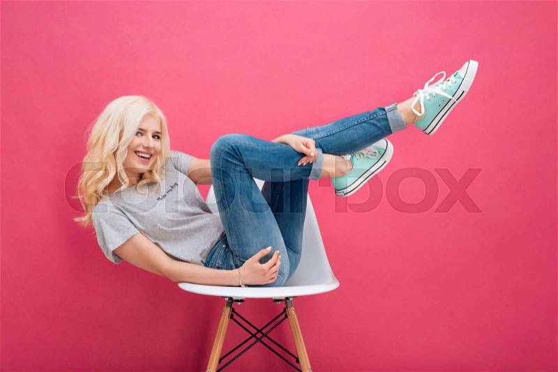 Smiling blonde woman sitting on the chair with raised legs over pink backgorund, stock photo