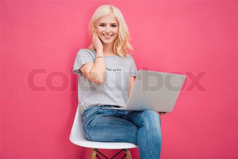 Attractive young woman sitting on the chair with laptop computer on pink background, stock photo