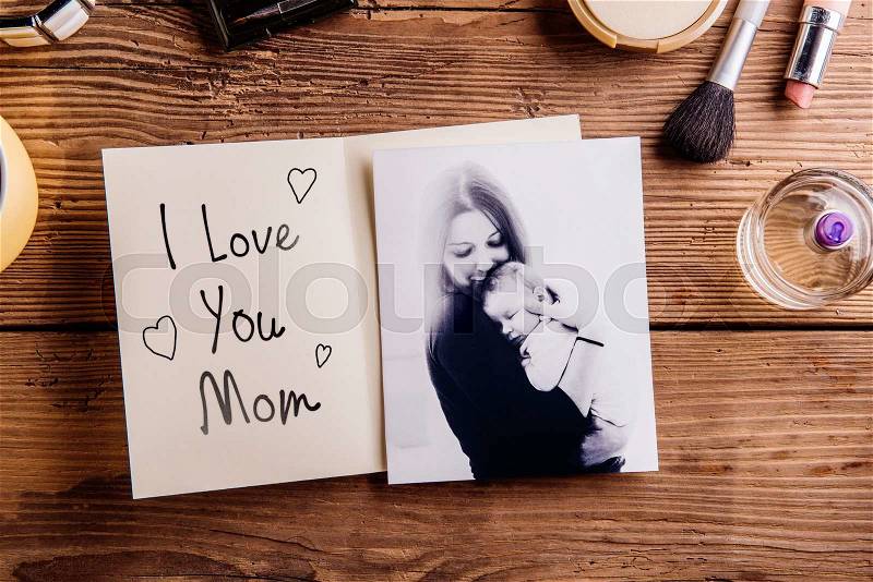 Mothers day composition. Black-and-white picture of mother holding her little baby, greeting card with I love you Mom text and various cosmetics. Studio shot on wooden background, stock photo