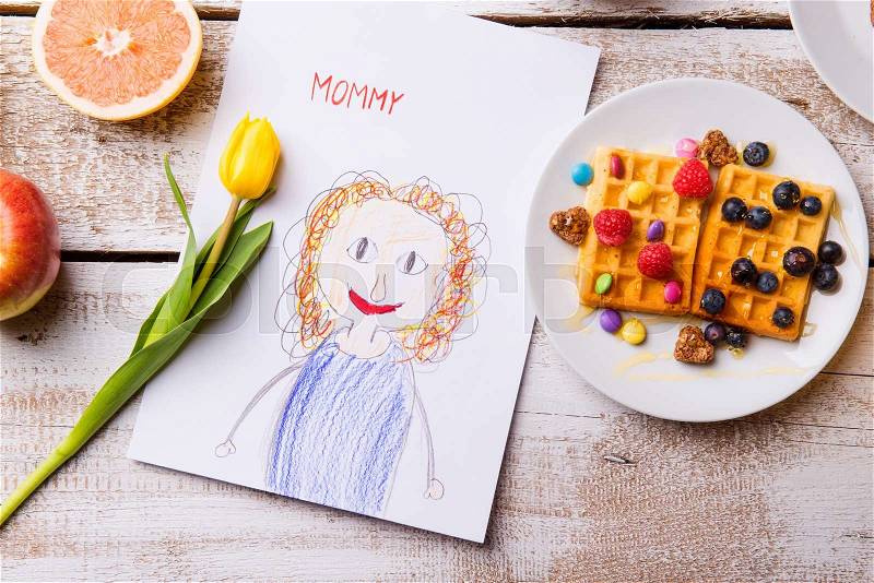 Mothers day composition. Childs drawing of her mother, yellow tulip and breakfast waffles with fruit. Studio shot on wooden background, stock photo
