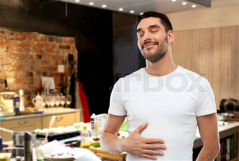 Eating, satisfaction and people concept - happy full man touching his tummy over kitchen background, stock photo