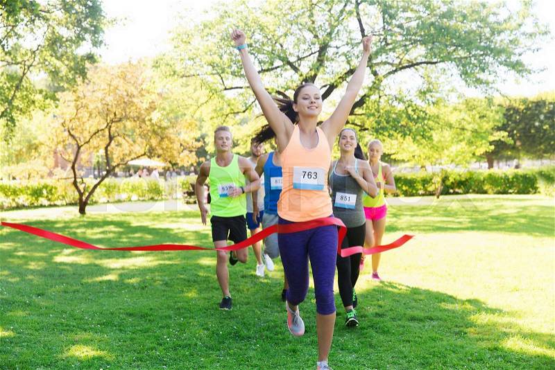 Fitness, sport, victory, success and healthy lifestyle concept - happy woman winning race and coming first to finish red ribbon over group of sportsmen running marathon with badge numbers outdoors, stock photo
